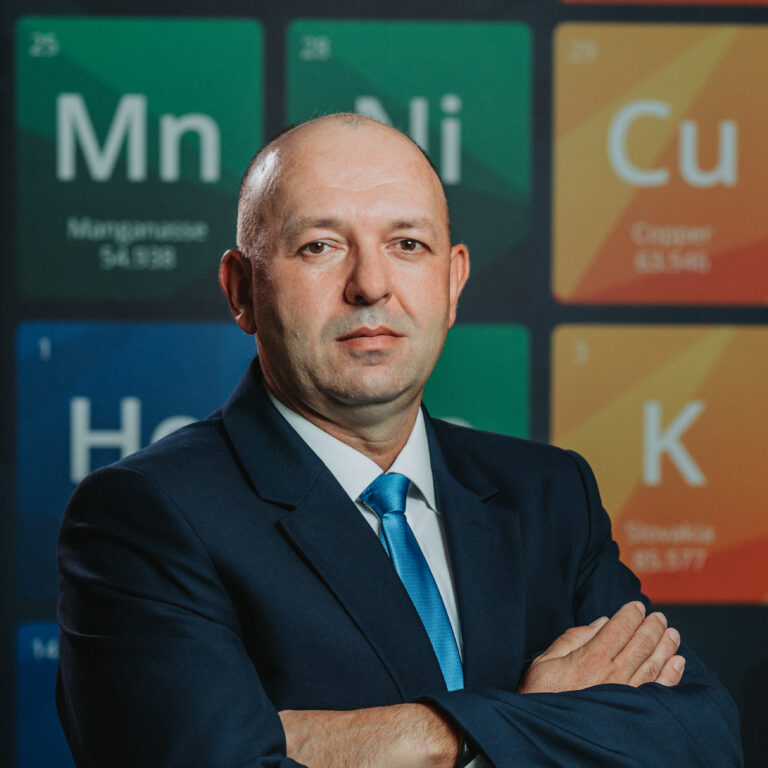 Muž v obleku s modrou kravatou stojí s prekríženými rukami pred stenou zobrazujúcou periodickú tabuľku prvkov, ktorá predstavuje záväzok spoločnosti Heneken Conductors k vedeckej dokonalosti.