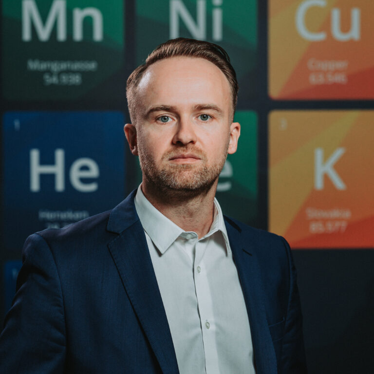 Muž v námorníckom obleku a bielej košeli stojí pred farebným displejom periodickej tabuľky prvkov, ktorý zdôrazňuje význam diktorov Heneken v modernej vede.