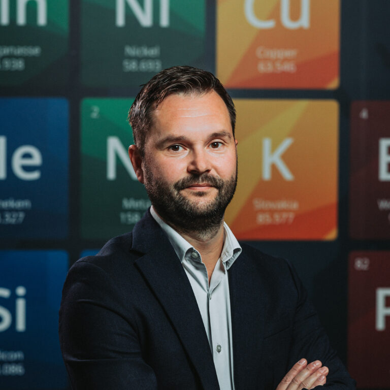 Muž v obleku stojí so skríženými rukami pred stenou s prvkami periodickej tabuľky, ktorá symbolizuje presnosť a odbornosť spoločnosti Heneken Conductors.